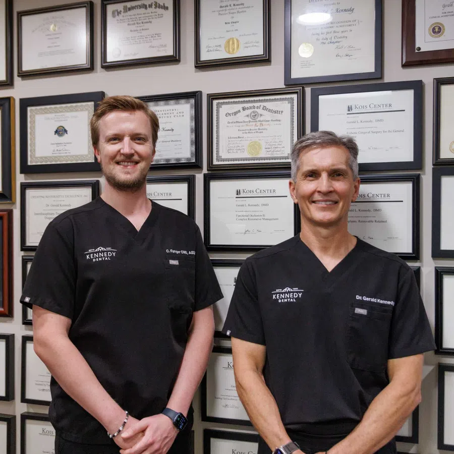 Two dentists wearing black Kennedy Dental uniforms stand outside the dental office under a Kennedy Dental sign.