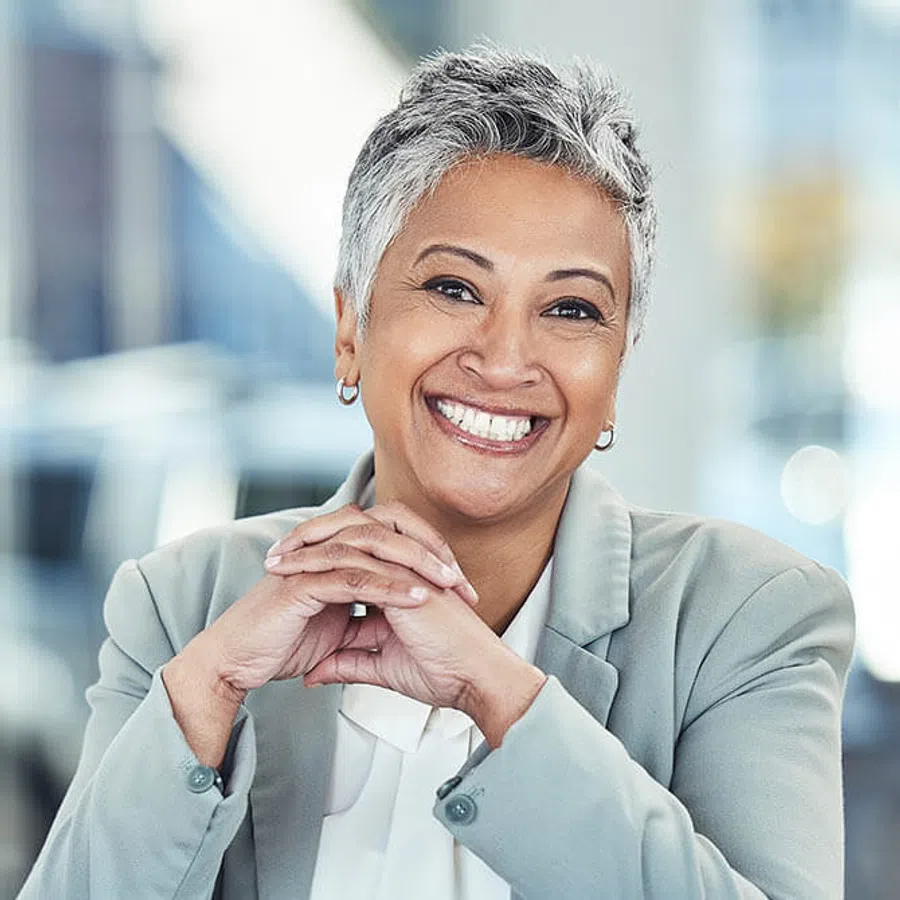 Smiling mature woman with gray hair in a light gray blazer, seated with hands clasped, blurred office background.