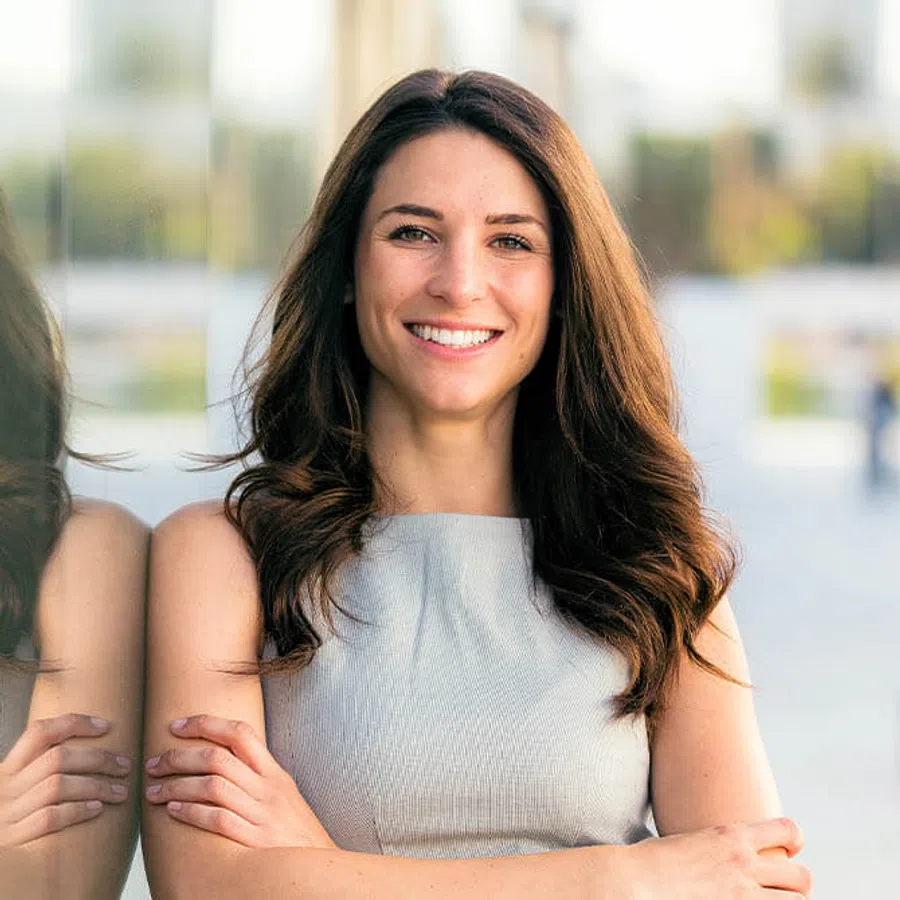 Confident young woman with long brown hair smiling with arms crossed, standing next to a reflective glass surface.
