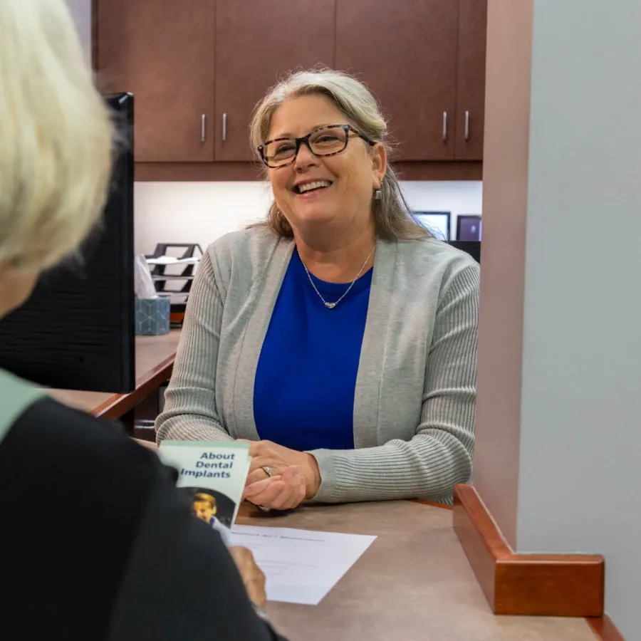 Smiling female dental professional consulting with a patient at a clinic reception desk with brochure about dental implants.
