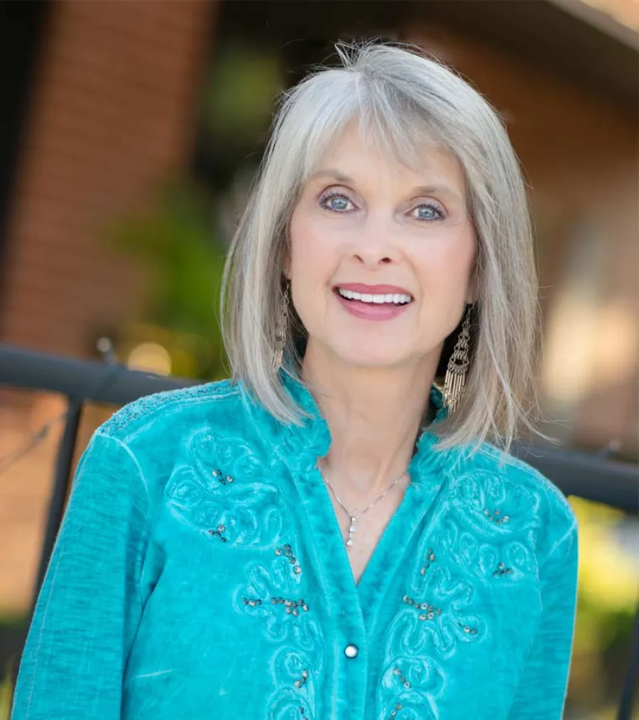 Smiling older woman with gray hair wearing a turquoise embroidered blouse and dangling earrings outdoors.