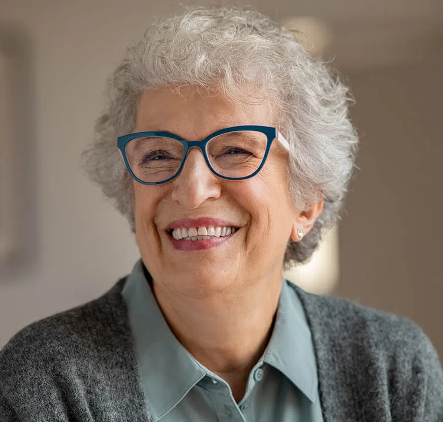 Smiling elderly woman with curly gray hair wearing blue glasses and a gray sweater in soft indoor lighting