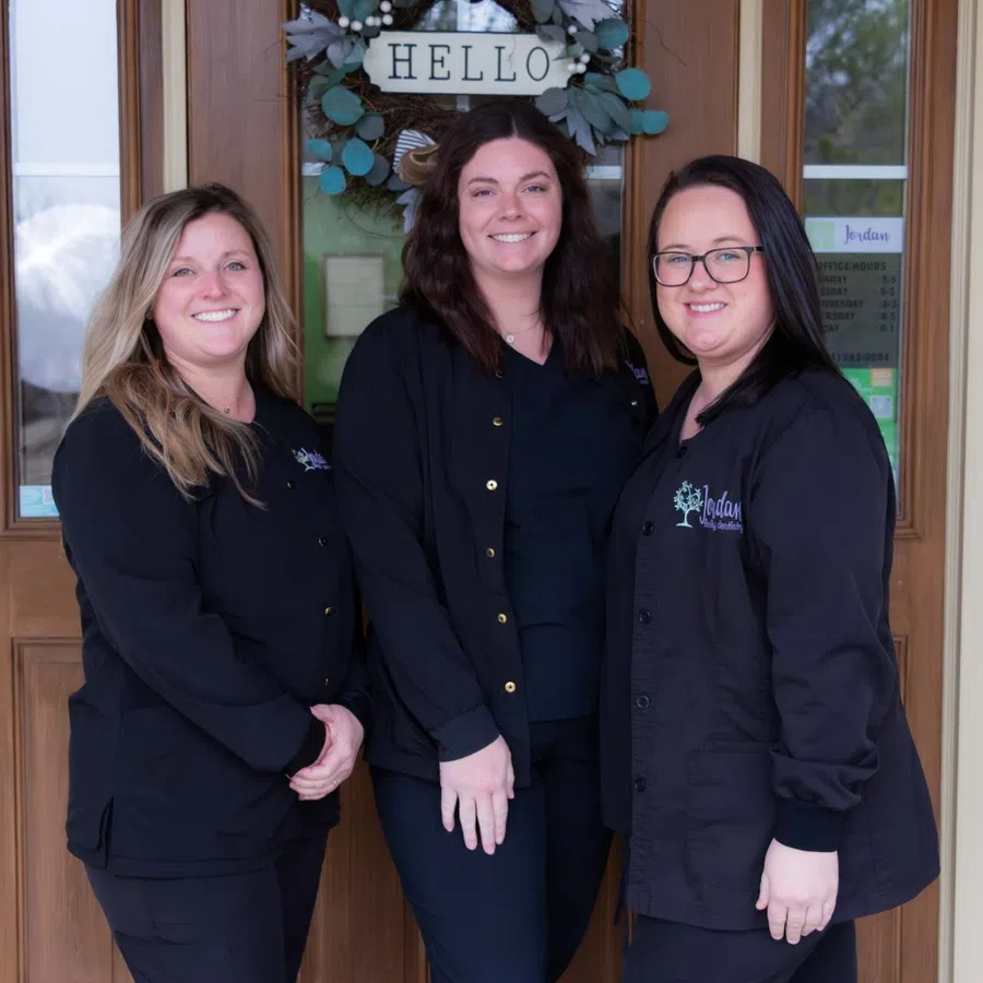 Three smiling women in black uniforms standing outside a door with a Hello sign and wreath