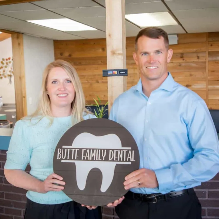 Two dental professionals holding a Butte Family Dental wooden sign inside a modern office with wood paneling.