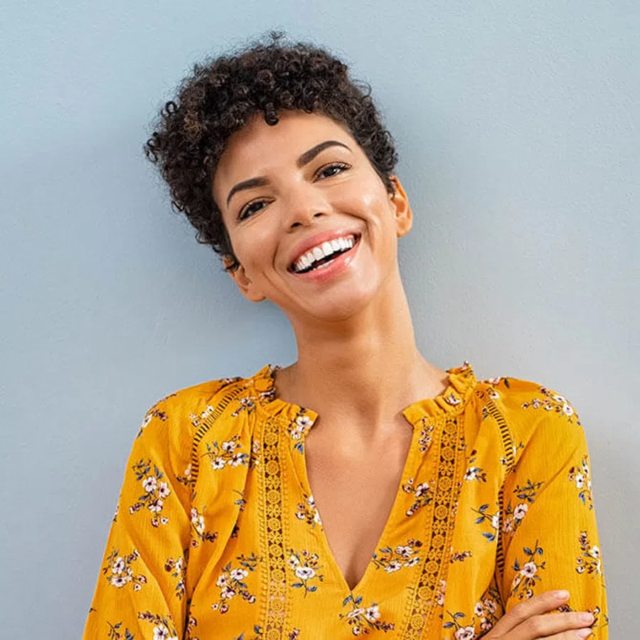 Smiling woman with short curly hair wearing a yellow floral blouse standing against a gray wall