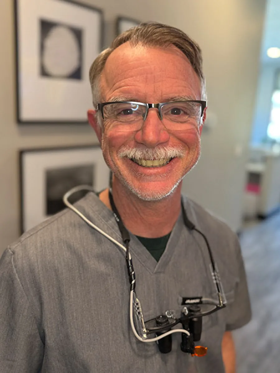 Smiling middle-aged male dentist wearing gray scrubs and magnifying glasses in a bright dental office.