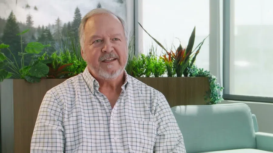 Smiling elderly man in a plaid shirt sitting in a bright room with green plants and large windows.