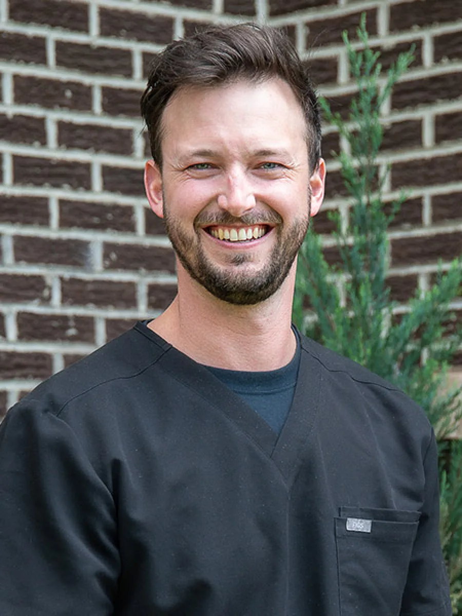 Smiling man with short brown hair and beard wearing black scrub top against brick wall and greenery background