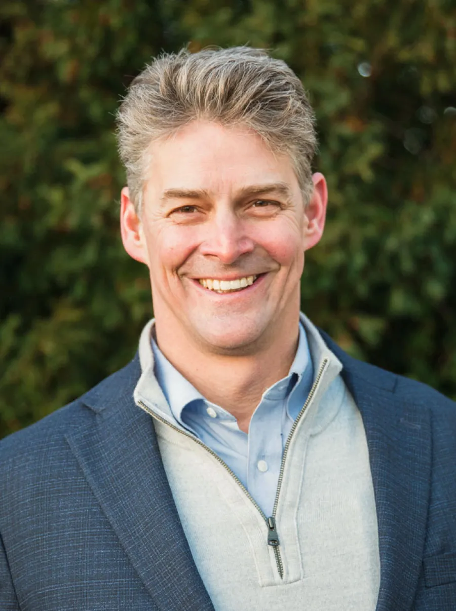 Smiling middle-aged man with gray hair wearing a blue blazer and light gray zip-up sweater outdoors.
