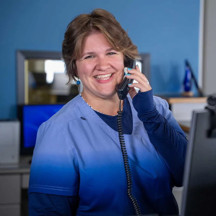 Smiling woman in blue scrubs talking on phone at an office desk with a computer monitor in the background