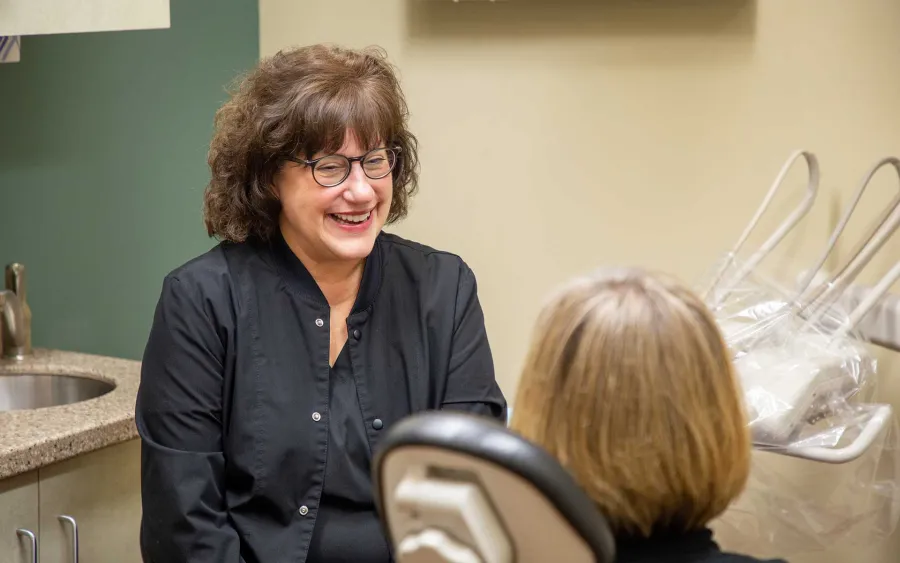 Smiling female dentist in black coat talking to female patient in dental office with equipment.