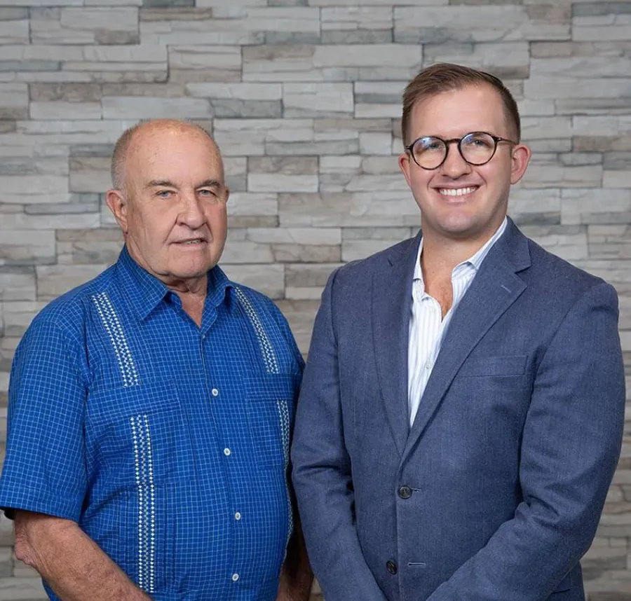Older man in blue shirt and younger man in suit stand smiling against stone wall background.