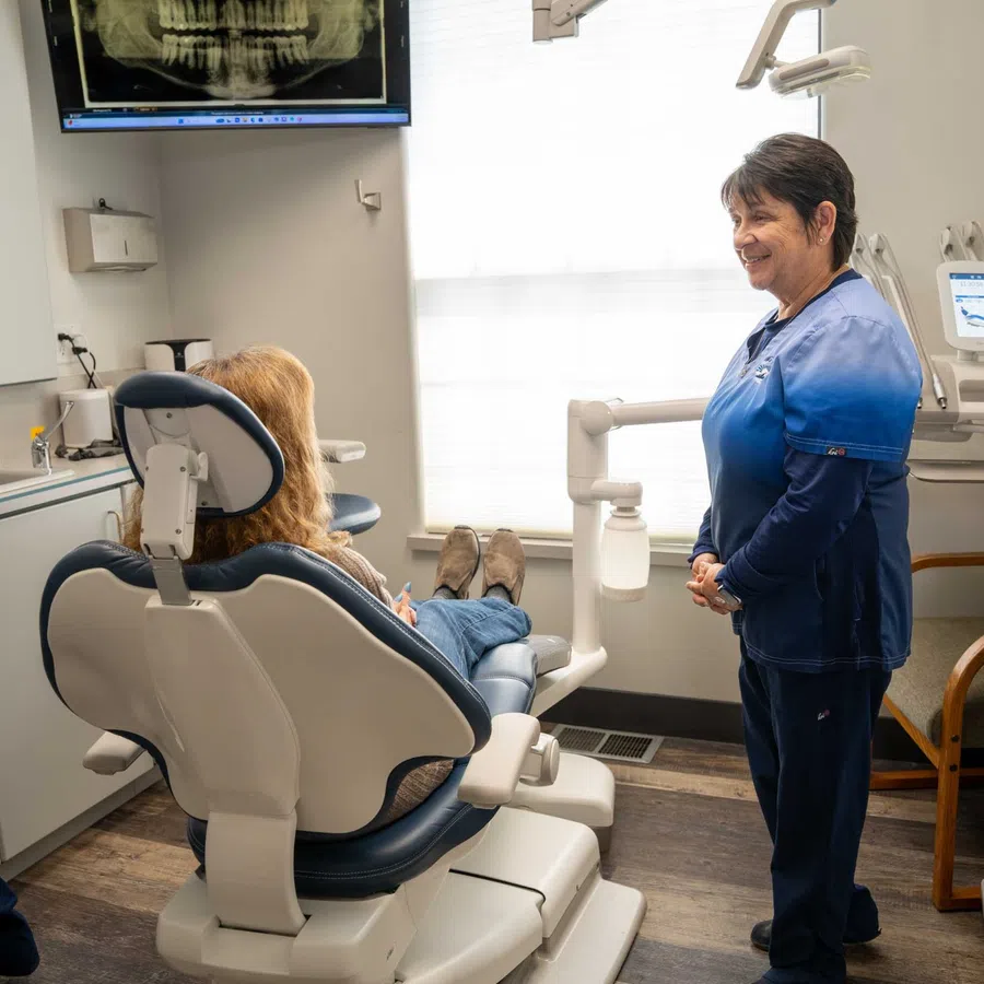 Dentist in blue scrubs smiling and talking to patient seated in dental chair with teeth X-ray on screen