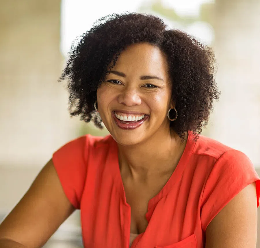 Smiling woman with curly hair wearing a red blouse and hoop earrings in a bright indoor setting