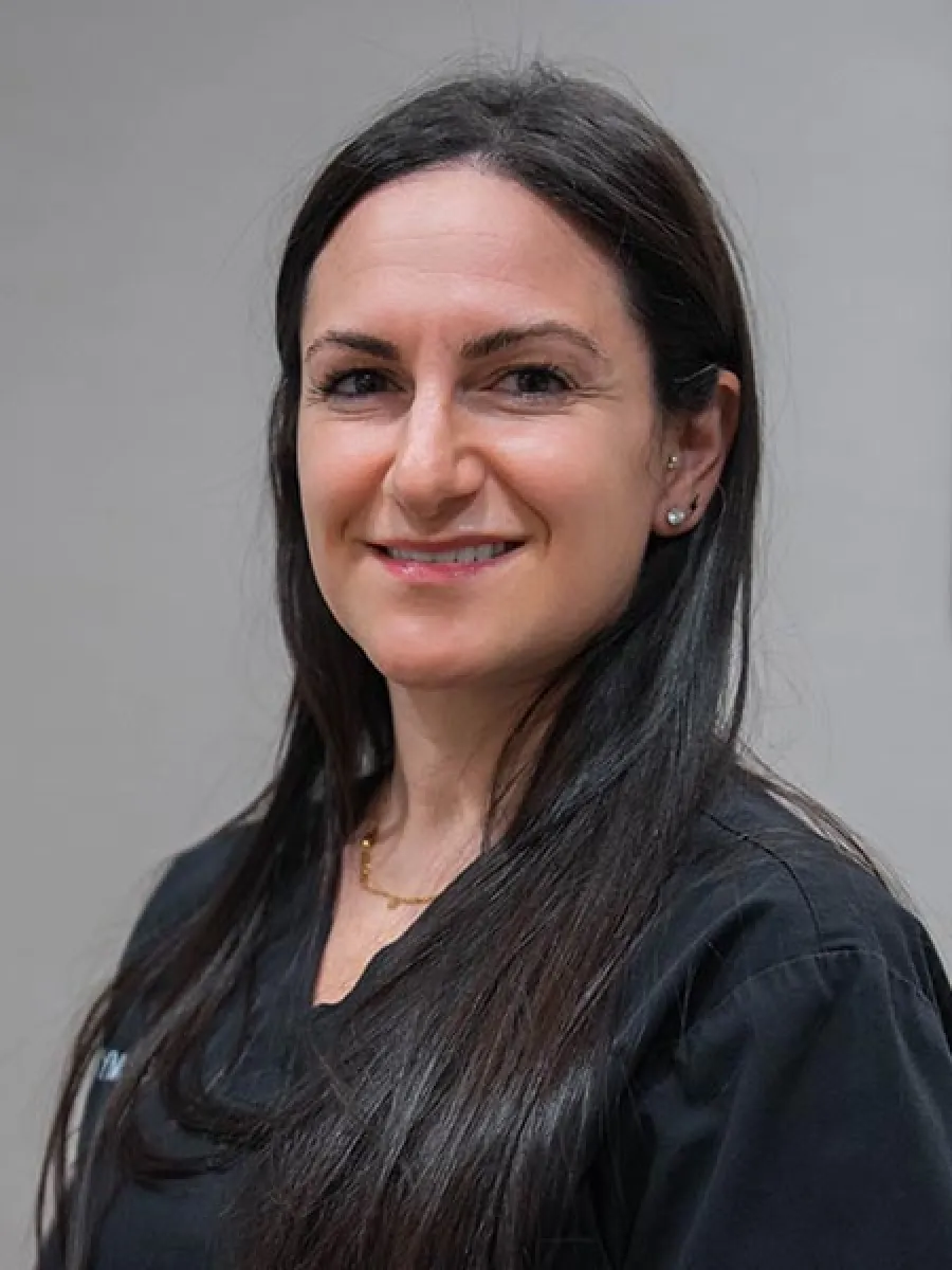 Smiling woman with long dark hair wearing black medical scrubs and stud earrings against a neutral background