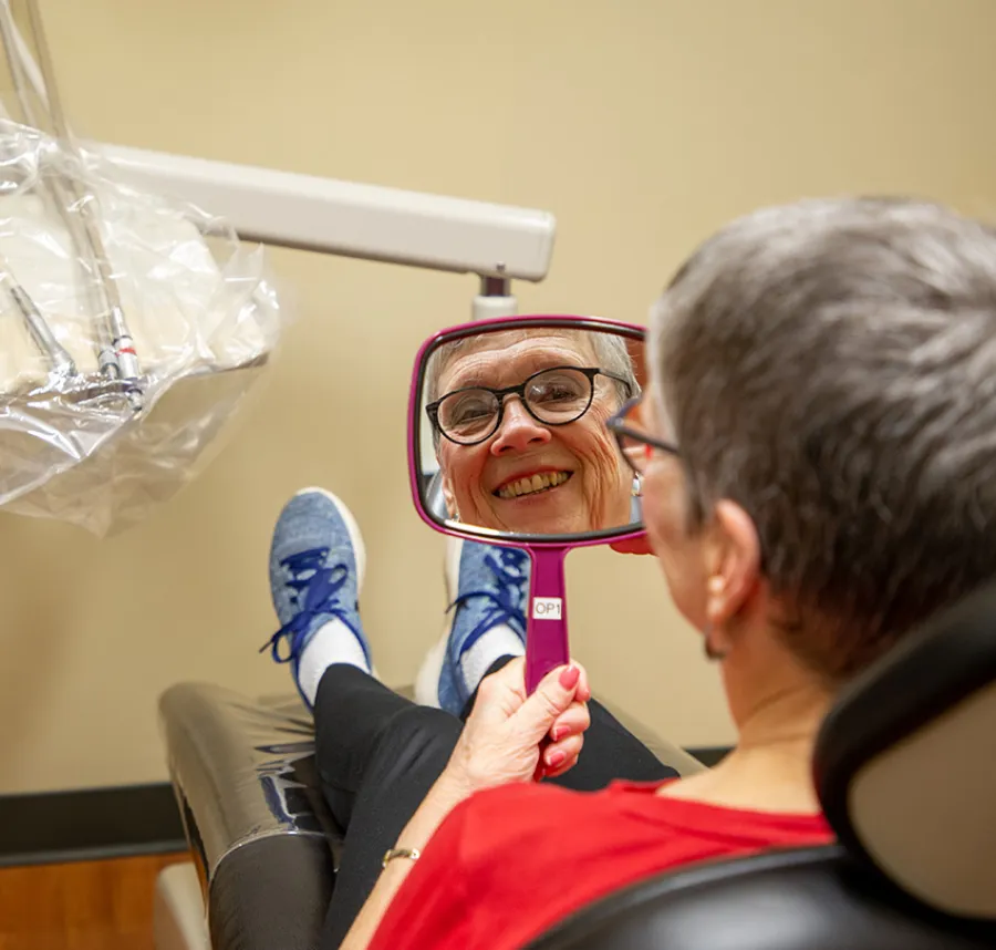 Elderly woman with glasses smiling at her reflection in a handheld mirror at a dental clinic.