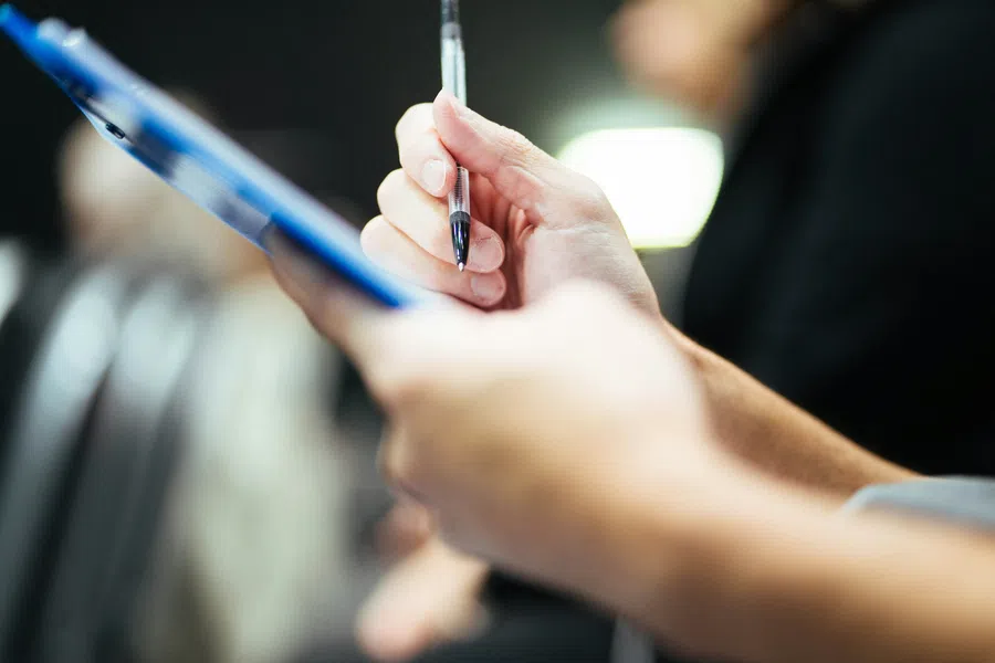 Person holding a pen and writing on a clipboard in a blurred indoor setting with a bright light in the background
