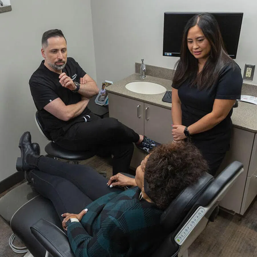 Patient reclining in a dental chair with two dental professionals consulting in a modern clinic room.