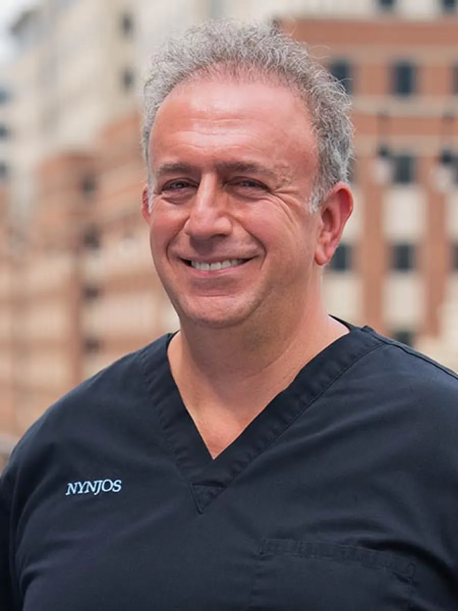 Smiling middle-aged man with gray hair wearing black medical scrubs with NYNJOS logo outdoors.