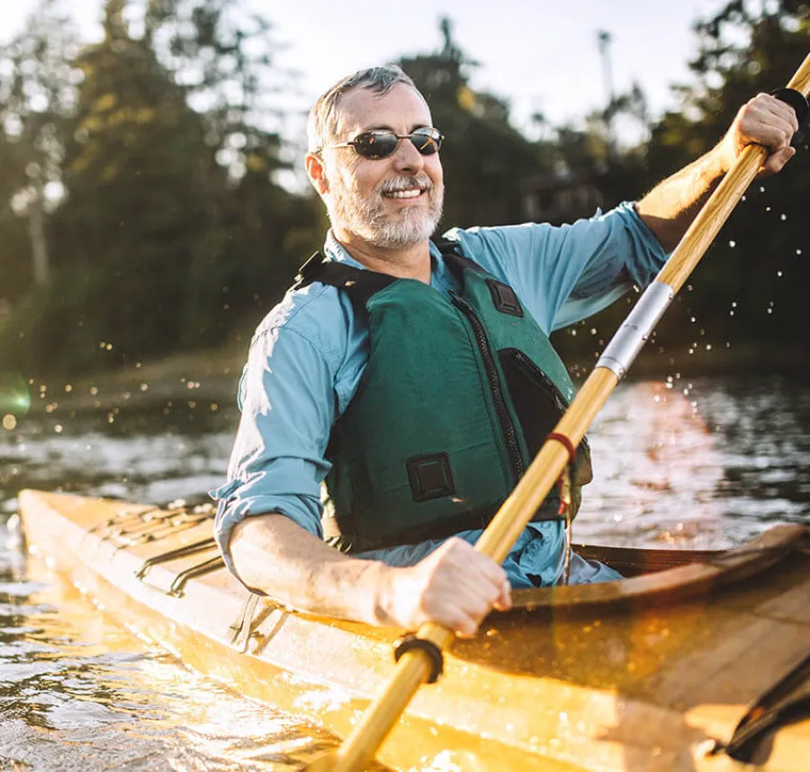 Smiling man wearing sunglasses and life jacket paddling a wooden kayak on a calm lake with trees in background