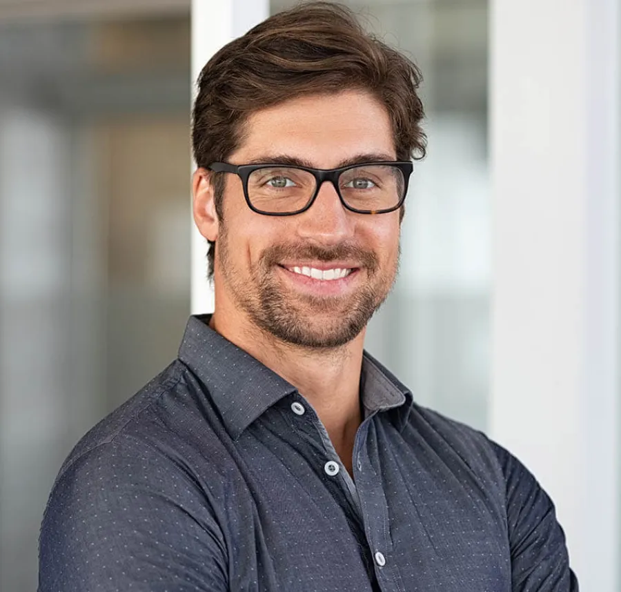 Smiling young man with glasses and beard wearing a dark button-up shirt in a modern office setting.