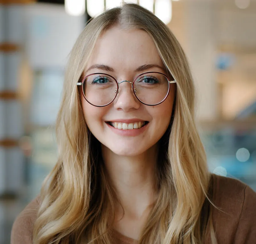 Young woman with long blonde hair wearing round glasses smiling indoors with blurred background.