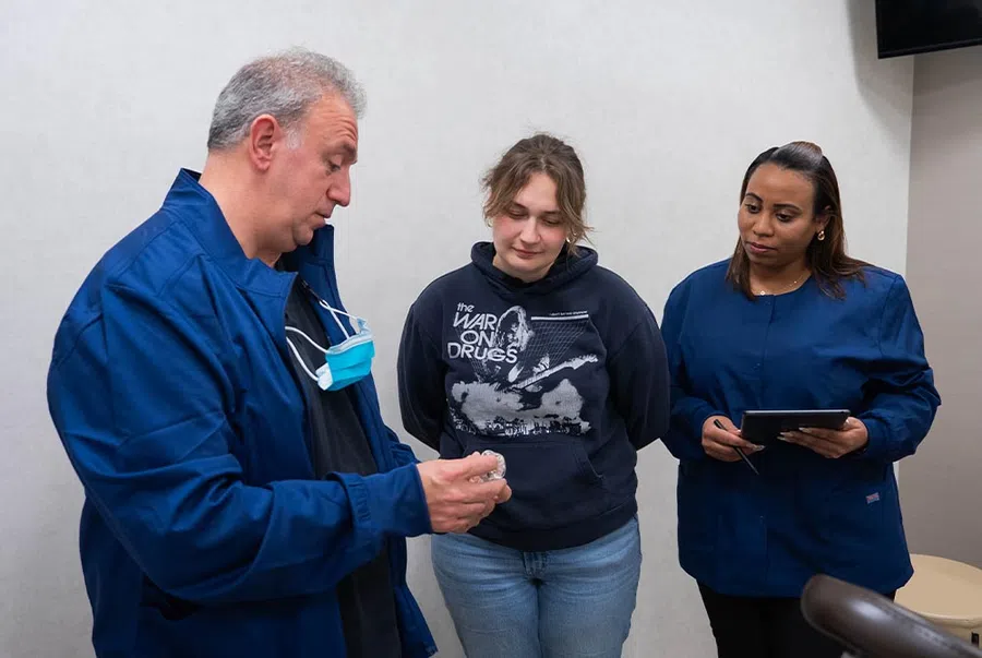 Three people engage in discussion indoors, one showing an object to two others, one holding a tablet.