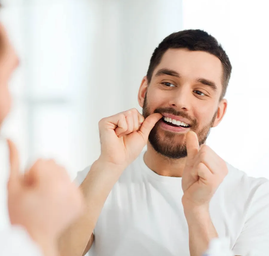 Man flossing his teeth in front of a bathroom mirror with a bright background, wearing a white shirt.
