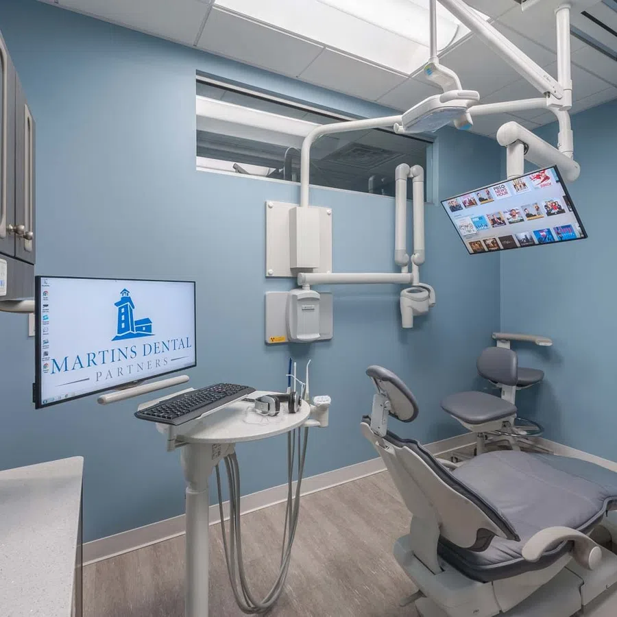 Modern dental treatment room with blue walls, dental chair, overhead monitor, and Martins Dental Partners logo on screen.