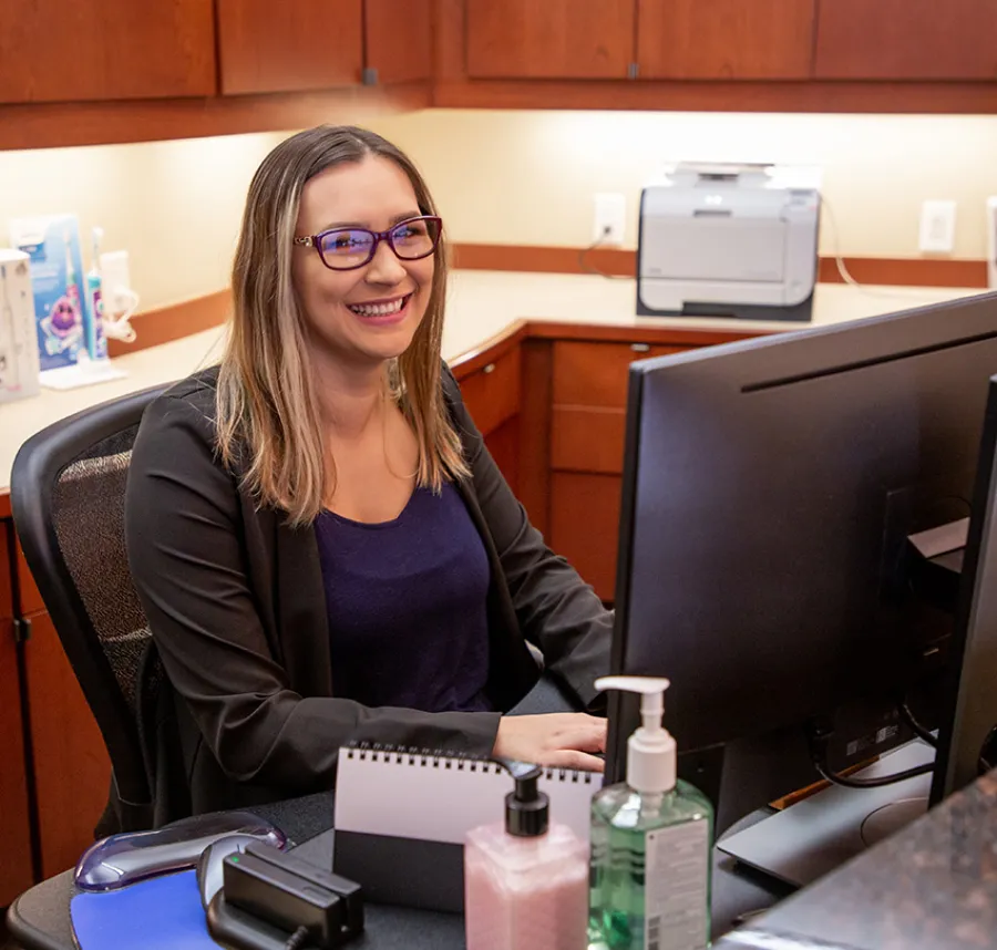 Smiling woman with glasses working at a computer in a modern office with wood cabinets and hand sanitizer bottles.
