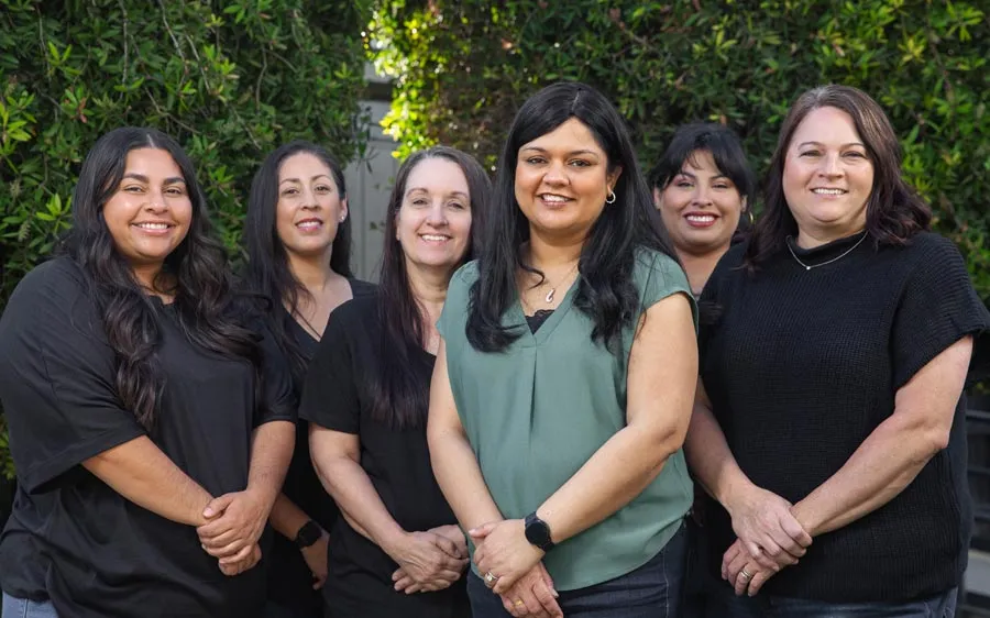 Diverse group of six women smiling outdoors with greenery background wearing casual clothing.