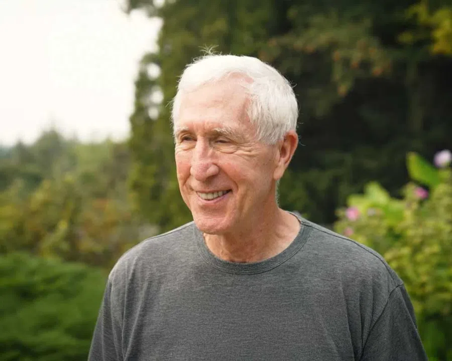 Elderly man with white hair smiling outdoors with blurred green foliage background