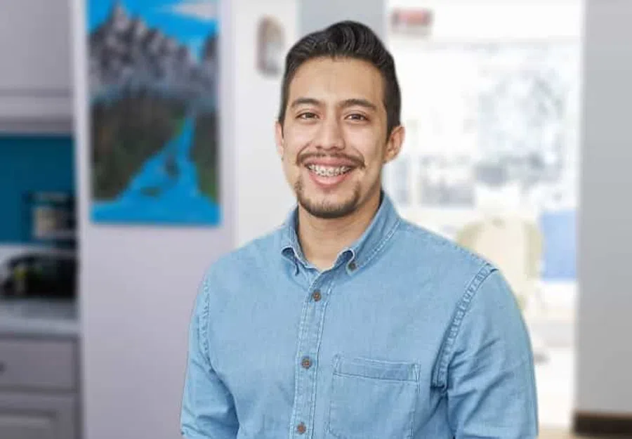 Smiling man with braces wearing a light blue denim shirt standing indoors with blurred background.