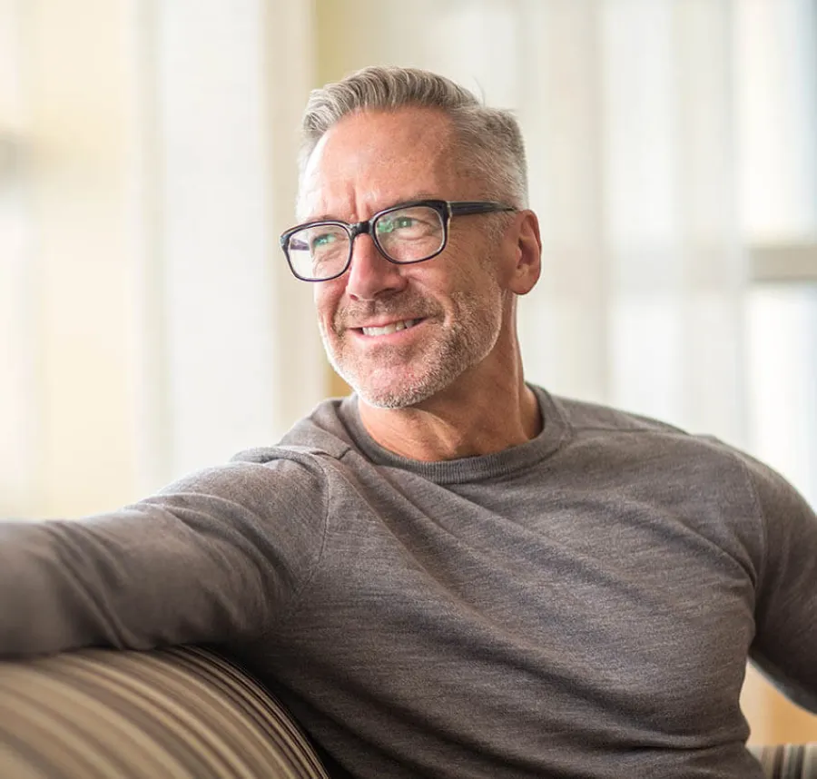 Middle-aged man with gray hair and glasses smiling while sitting on a striped couch in bright room.
