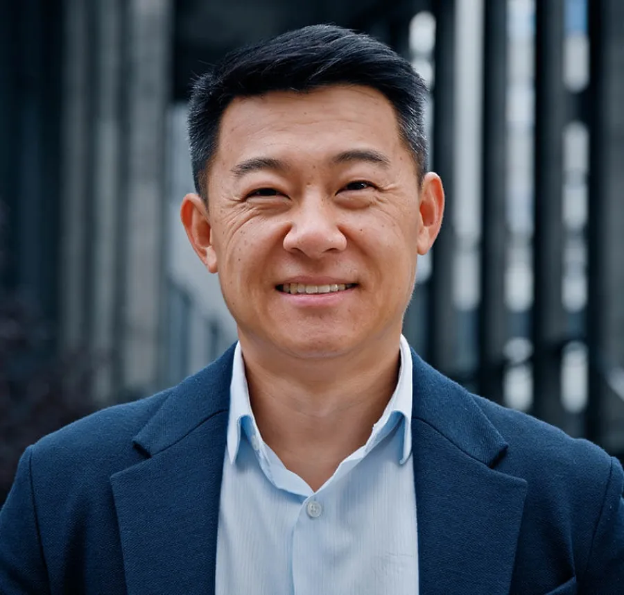 Smiling middle-aged Asian man wearing a navy blazer and light blue shirt in an urban outdoor setting.