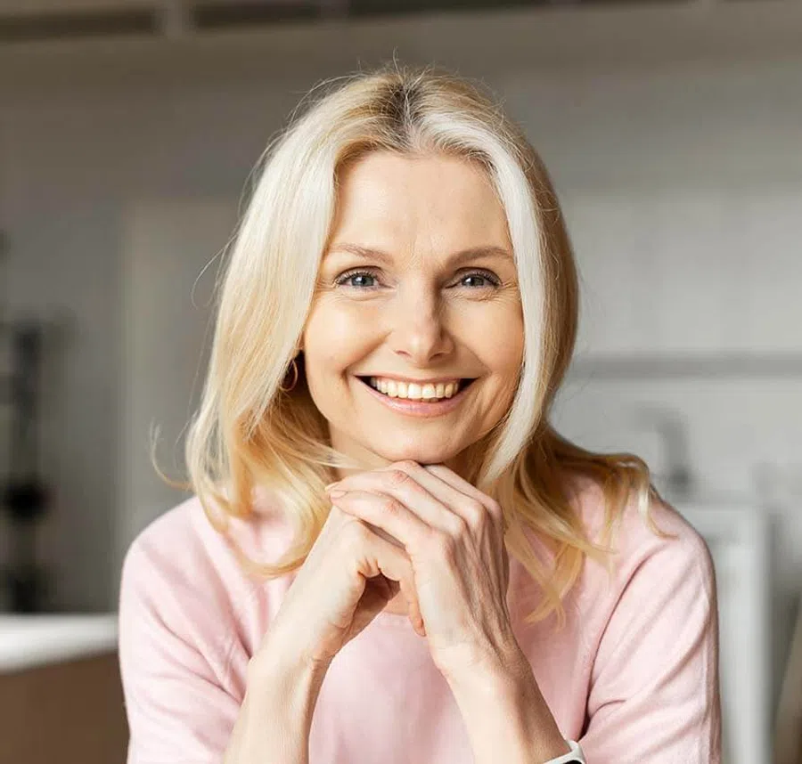 Smiling middle-aged woman with blonde hair wearing a light pink sweater, resting her chin on her hands indoors.