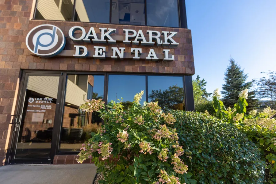 Exterior view of Oak Park Dental office with signage, glass door, and flowering plants under clear blue sky.