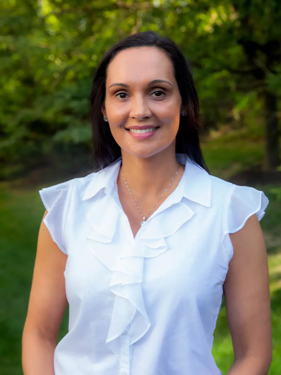 Smiling woman in white ruffled blouse outdoors with green trees in the background, natural lighting.