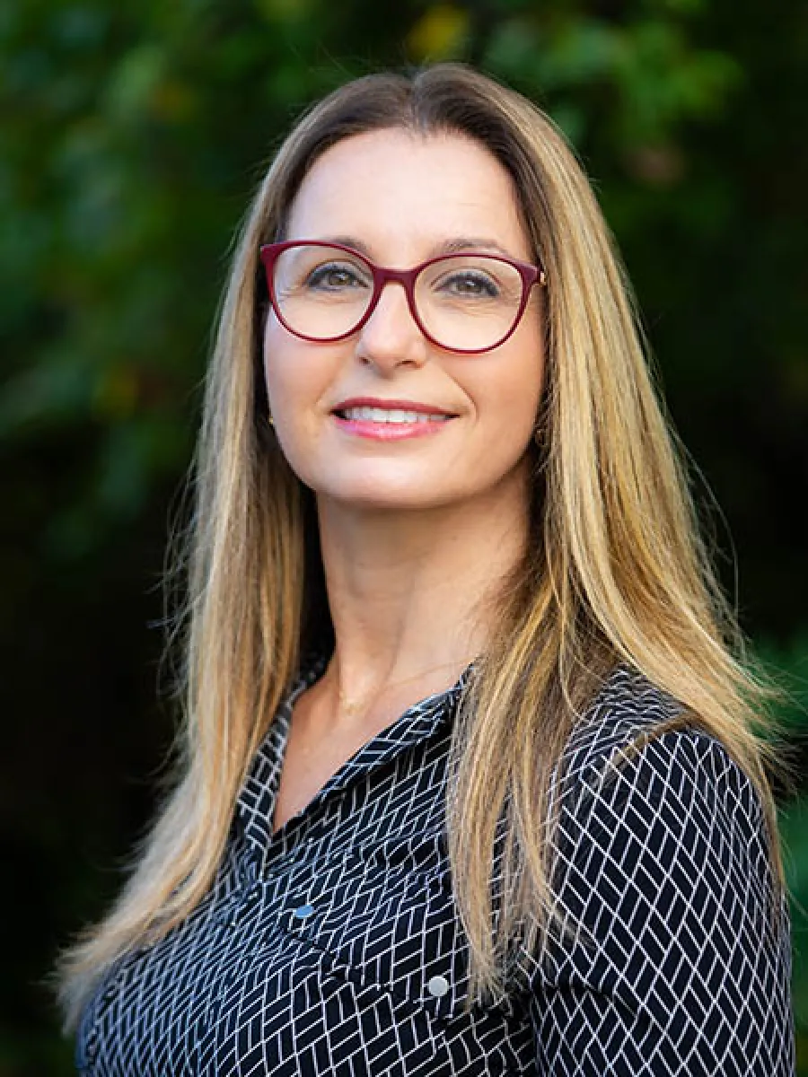 Smiling woman with long blonde hair wearing red glasses and a black patterned shirt outdoors.