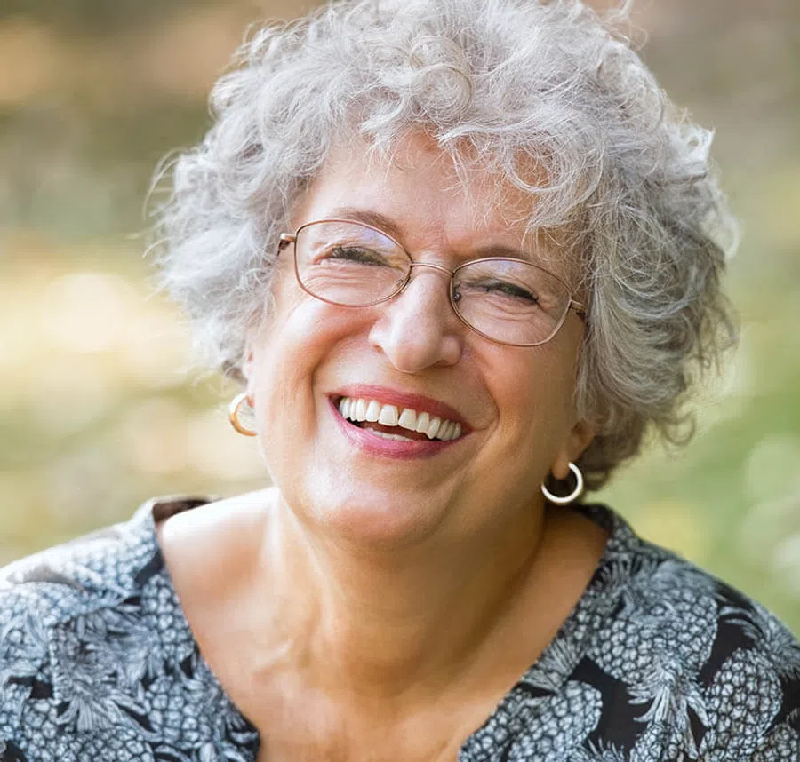 Elderly woman with curly gray hair and glasses smiling warmly outdoors in soft daylight.