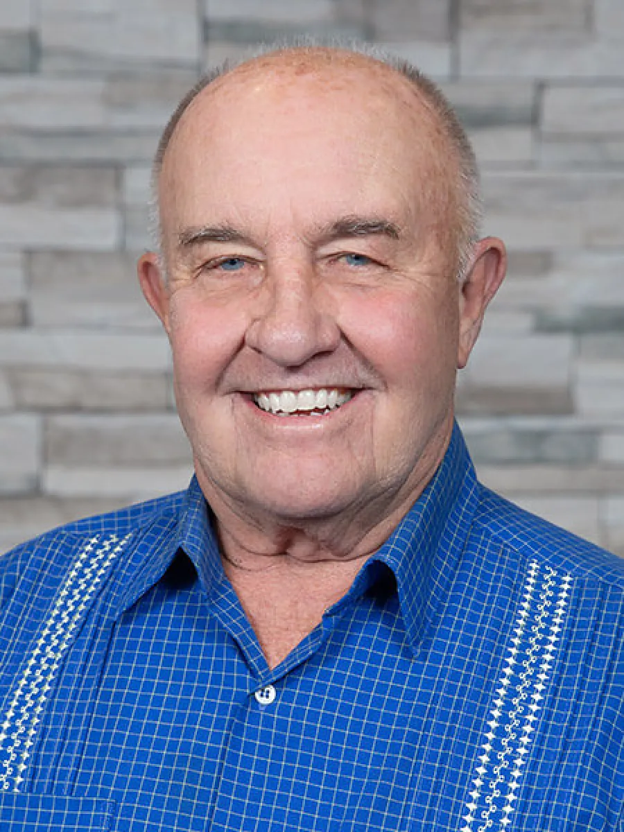 Smiling elderly man wearing a blue checkered shirt with a stone wall background.