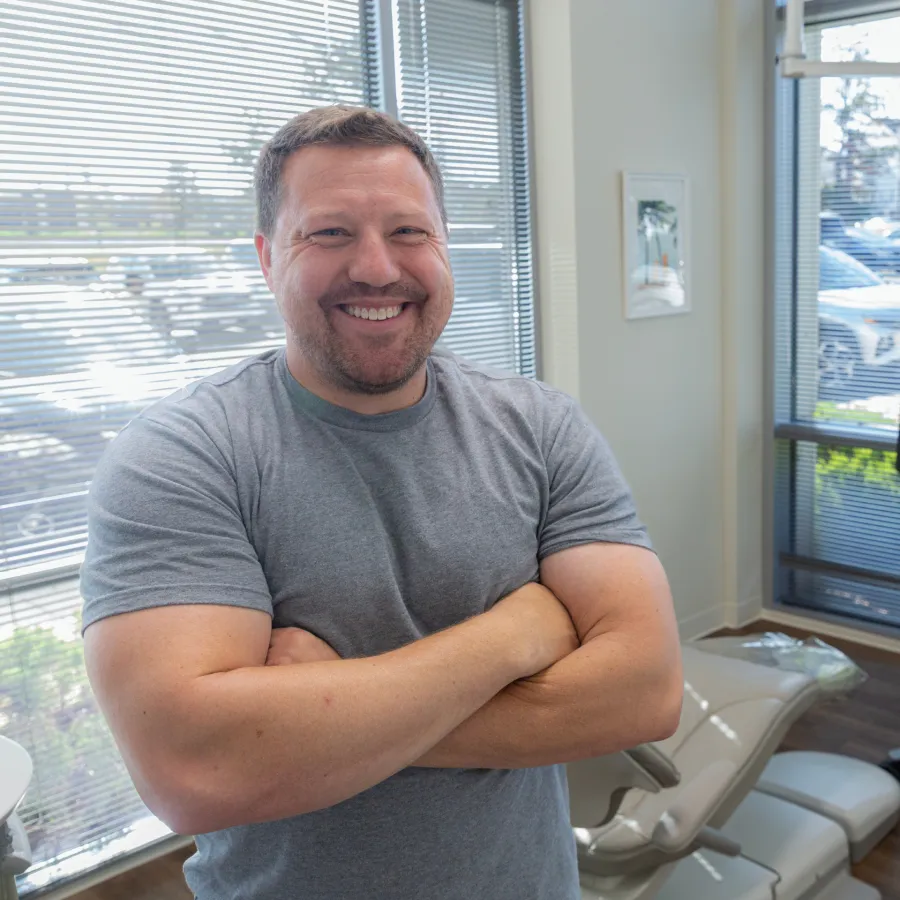 Smiling man with crossed arms wearing a gray t-shirt stands in a bright modern office with window blinds.