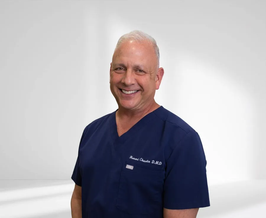 Smiling middle-aged male doctor wearing navy blue scrubs with white hair and a clean background.