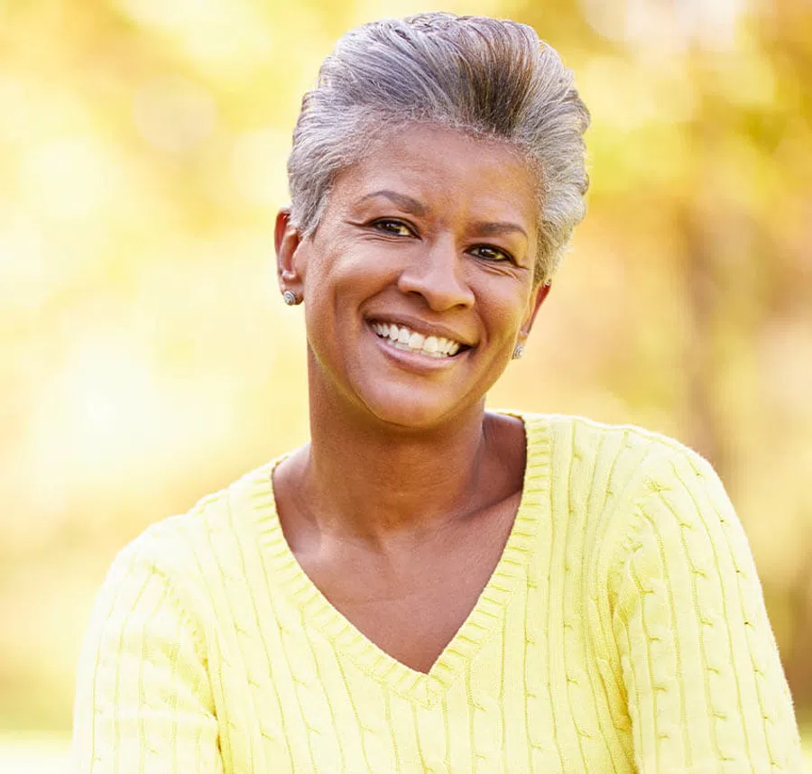 Smiling mature woman in yellow sweater with short gray hair against a soft natural background.