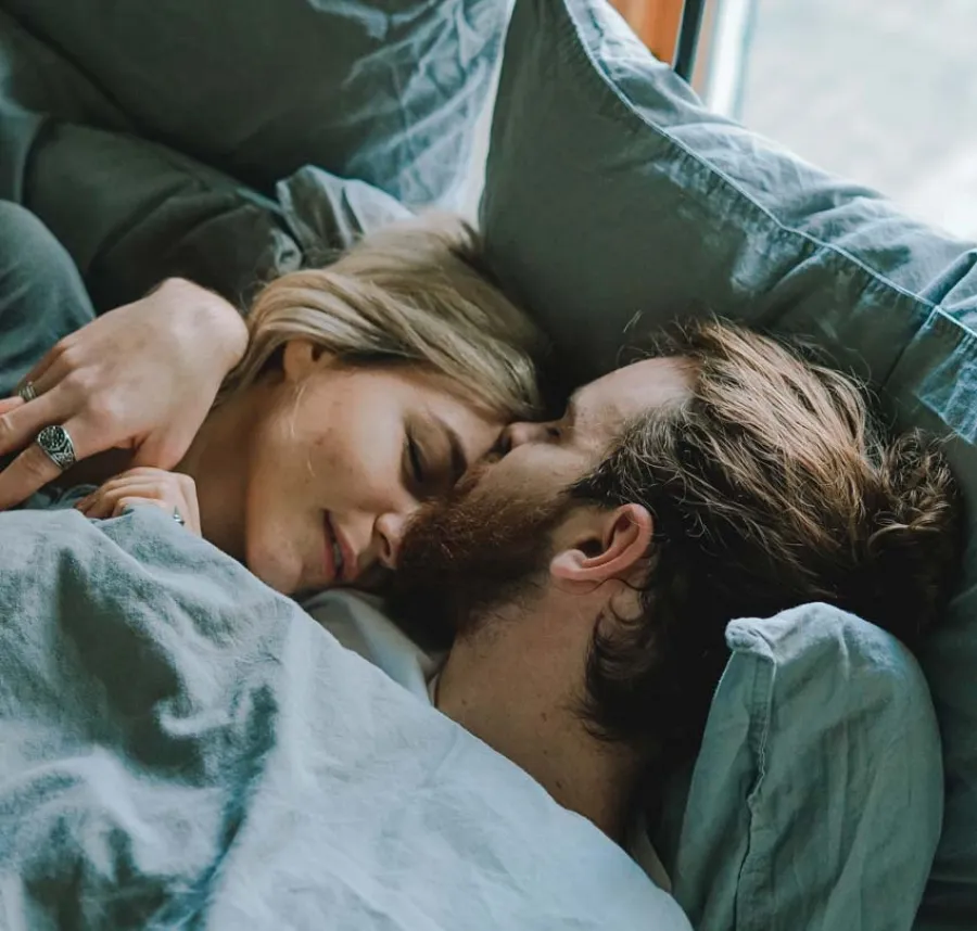 Couple cuddling closely and peacefully asleep together under blue bedding in a cozy bed by the window.