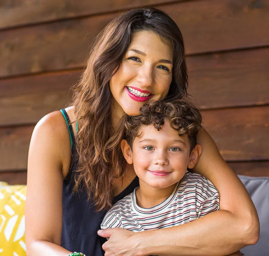 Smiling woman hugging young boy with curly hair sitting on couch against wooden wall background