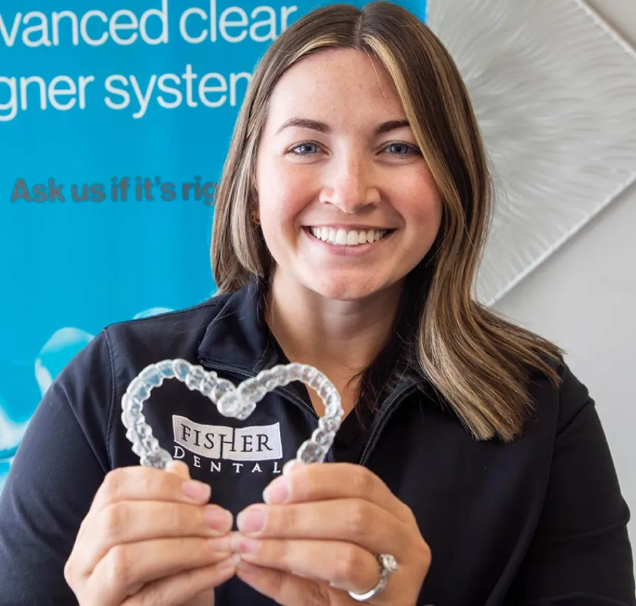 Smiling woman holding a heart-shaped clear dental aligner with dental office background.