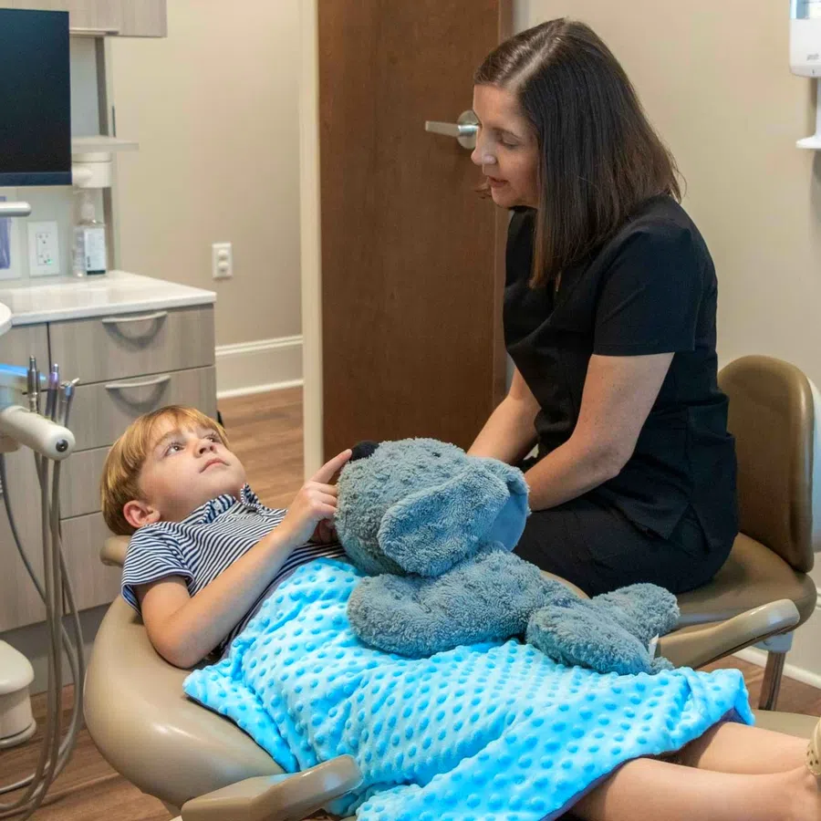 Child patient lying on dental chair with blue blanket and stuffed elephant toy, talking to female dentist in black scrubs