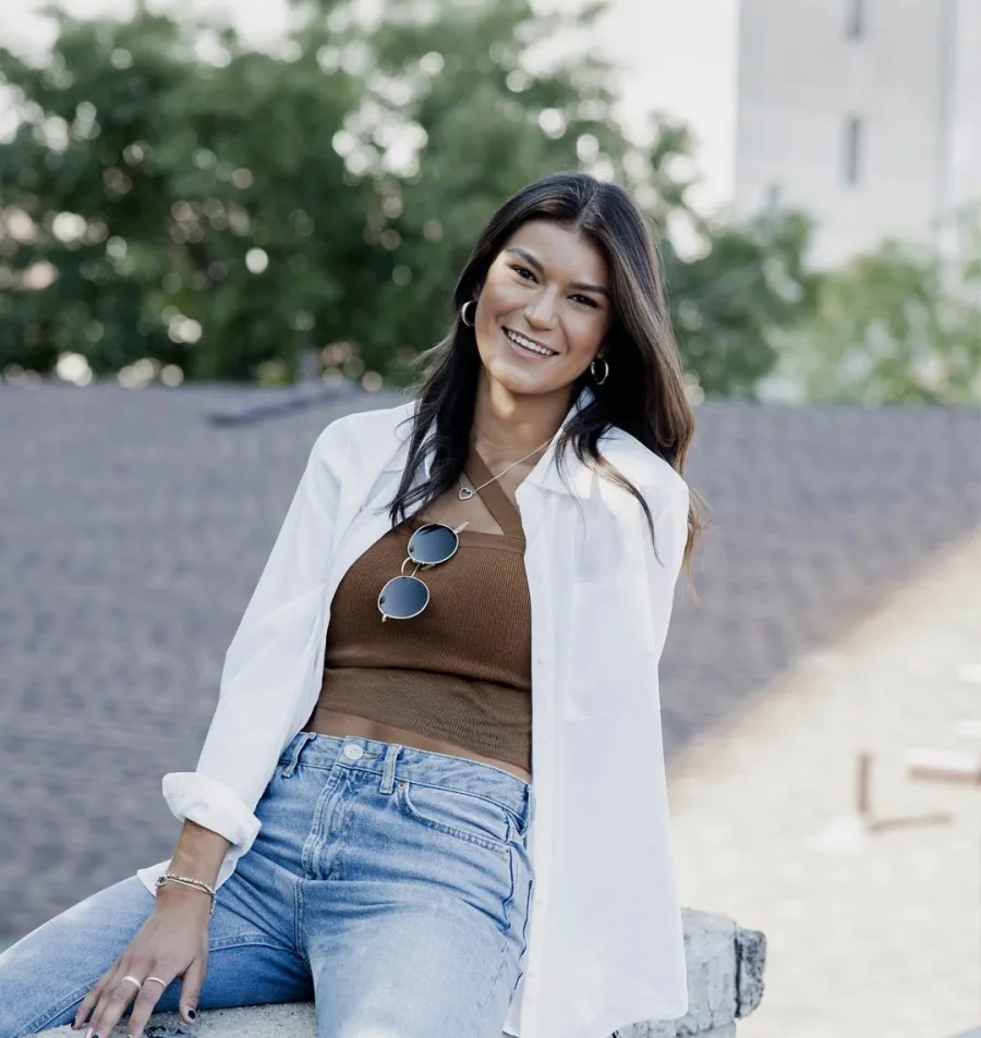 Smiling young woman wearing a brown tank top and white shirt leaning on a stone ledge outdoors