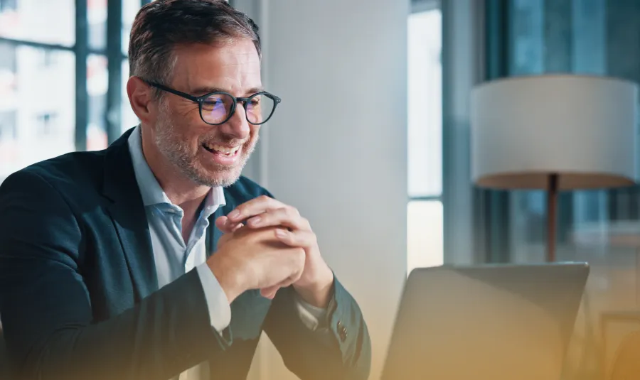 Smiling man in glasses and suit video chatting on laptop in a bright office with natural light and lamp.